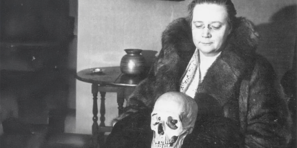 Archival black and white photograph of a woman in a fur coat seated with a human skull on her lap