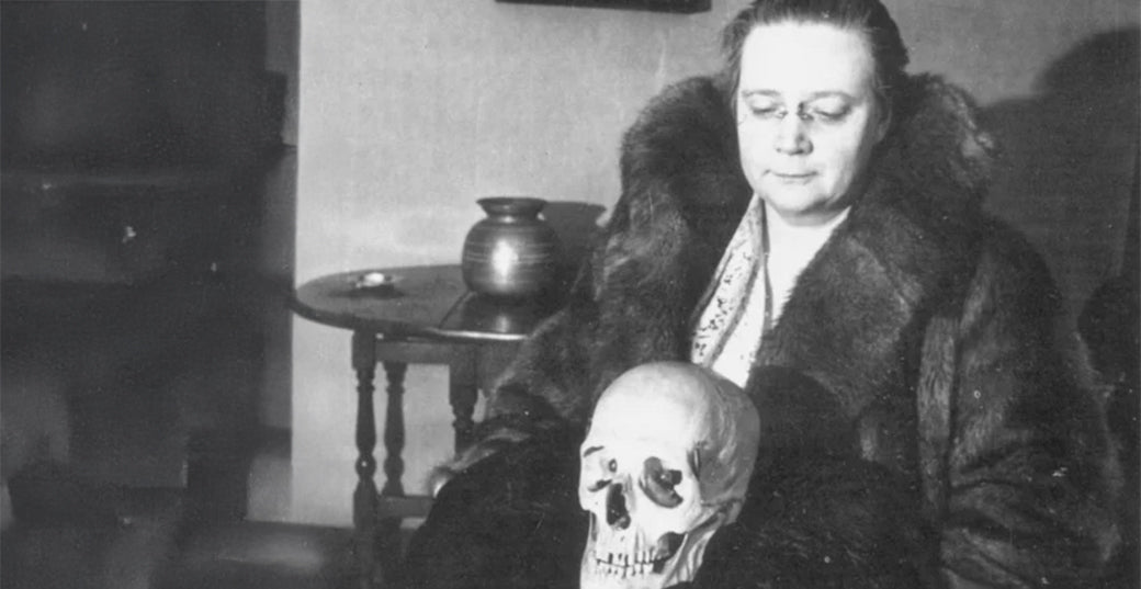 Archival black and white photograph of a woman in a fur coat seated with a human skull on her lap