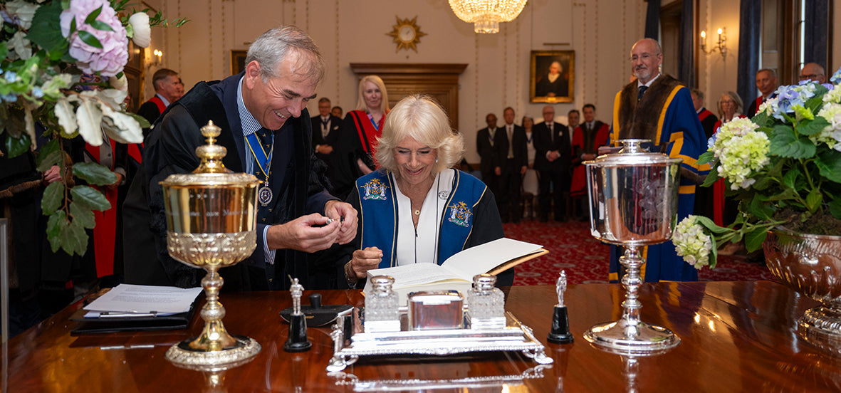 Photograph of Queen Camilla signing a document at a ceremonial desk with silver pen stands, flanked by officials