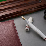 Estilográfica Conway Stewart Raleigh White Crystal apoyada sobre un escritorio junto a un bote de tinta y un soporte para plumas