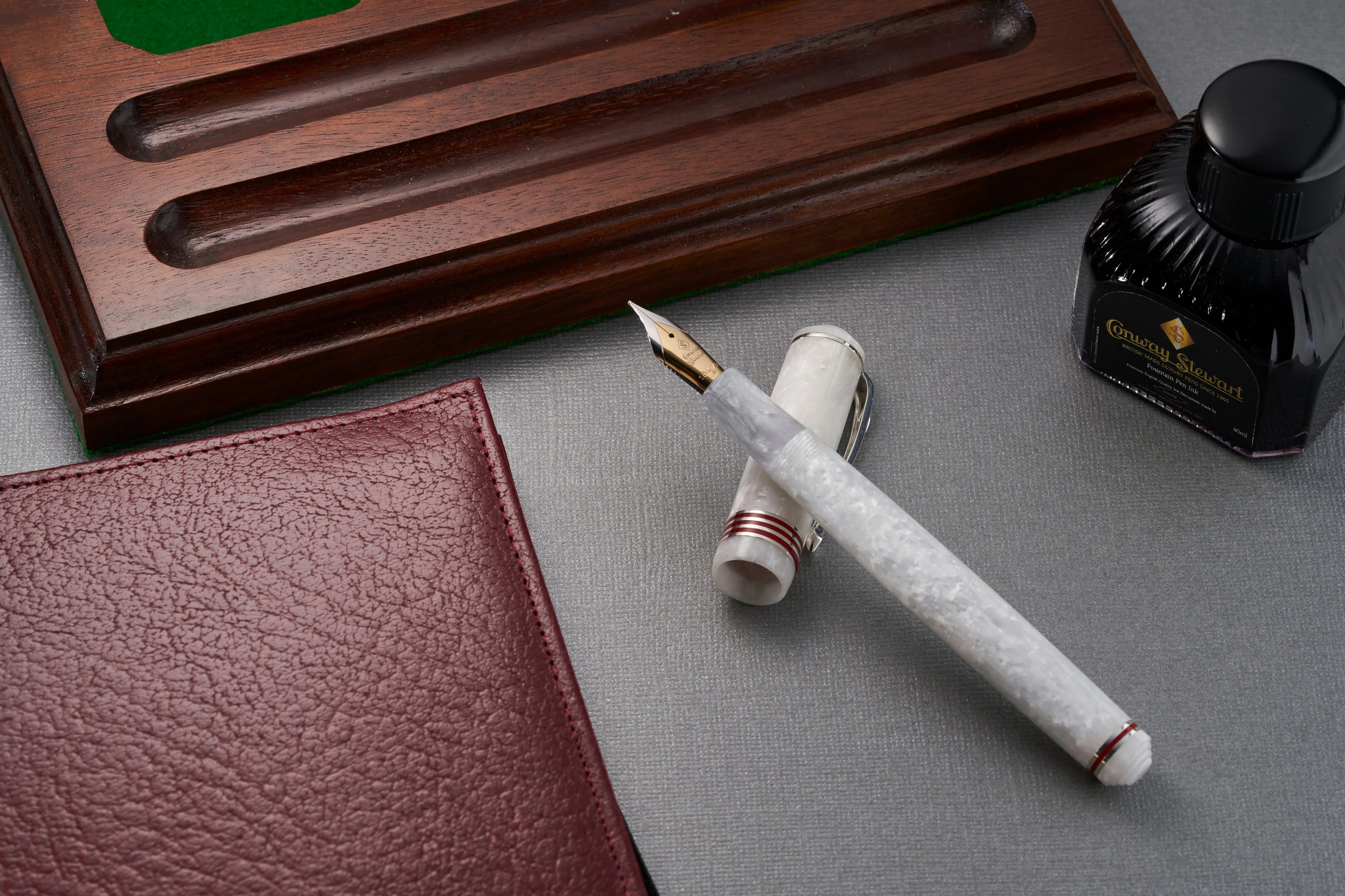 Estilográfica Conway Stewart Raleigh White Crystal apoyada sobre un escritorio junto a un bote de tinta y un soporte para plumas