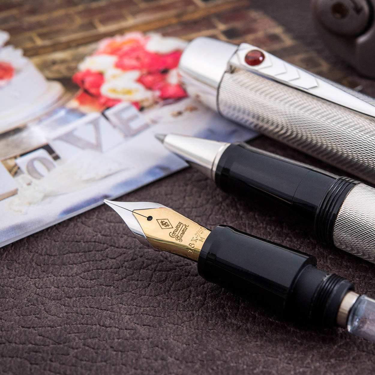 Pluma estilográfica de boda plateada con patrón de grano de cebada y joya de cabujón de rubí, apoyada junto a fotografías de boda