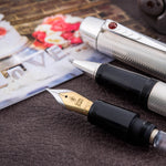 Pluma estilográfica de boda plateada con patrón de grano de cebada y joya de cabujón de rubí, apoyada junto a fotografías de boda