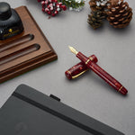 Winston Cherry Red Füllfederhalter über Kreuz auf einem grauen Schreibtisch, mit Tintenflasche, Tannenzapfen und Winterbeeren