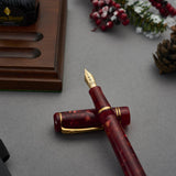 Winston Cherry Red Füllfederhalter über Kreuz neben einem geschlossenen Notizbuch, mit Beeren und Tannenzapfen im Hintergrund