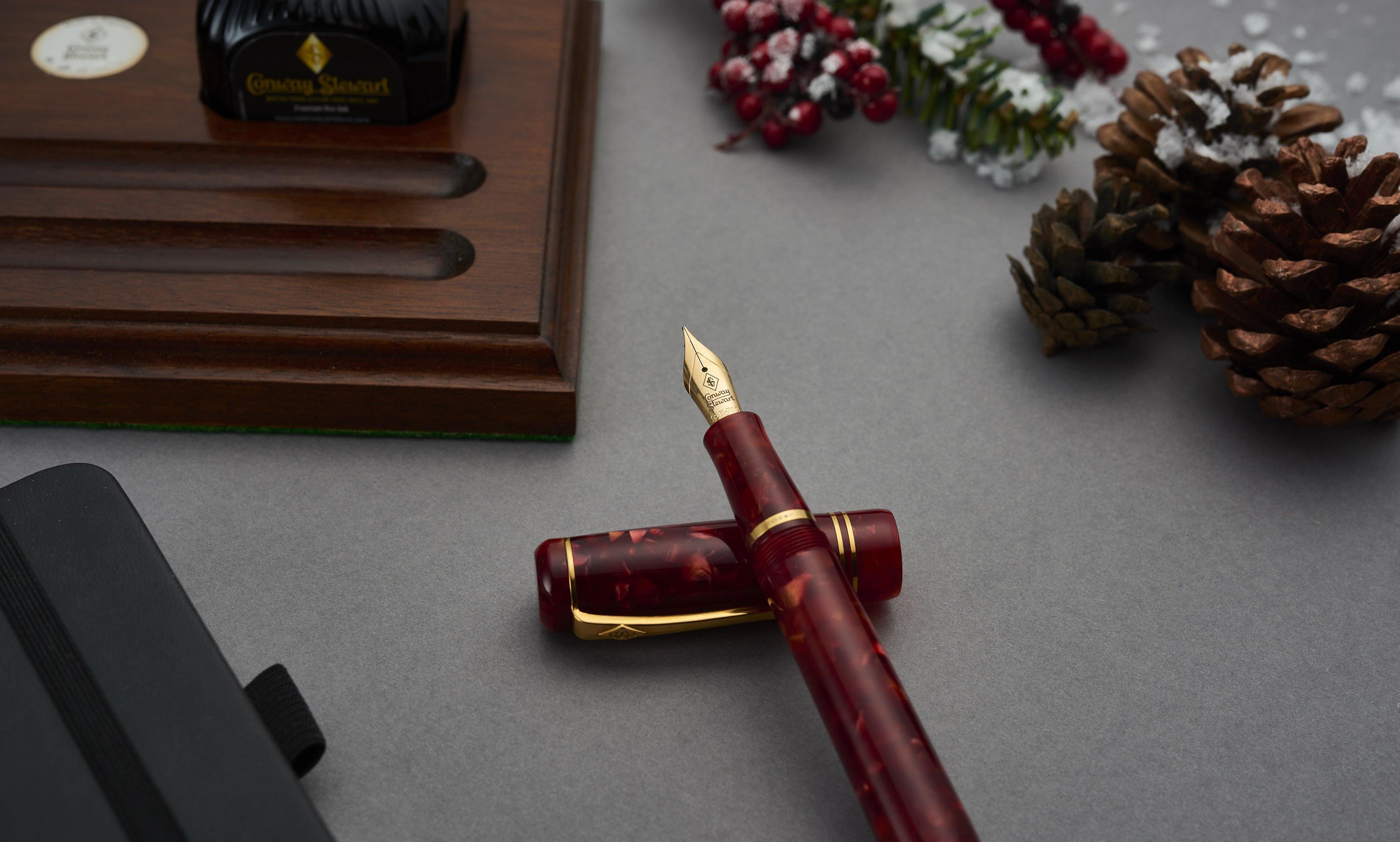 Winston Cherry Red Füllfederhalter über Kreuz neben einem geschlossenen Notizbuch, mit Beeren und Tannenzapfen im Hintergrund